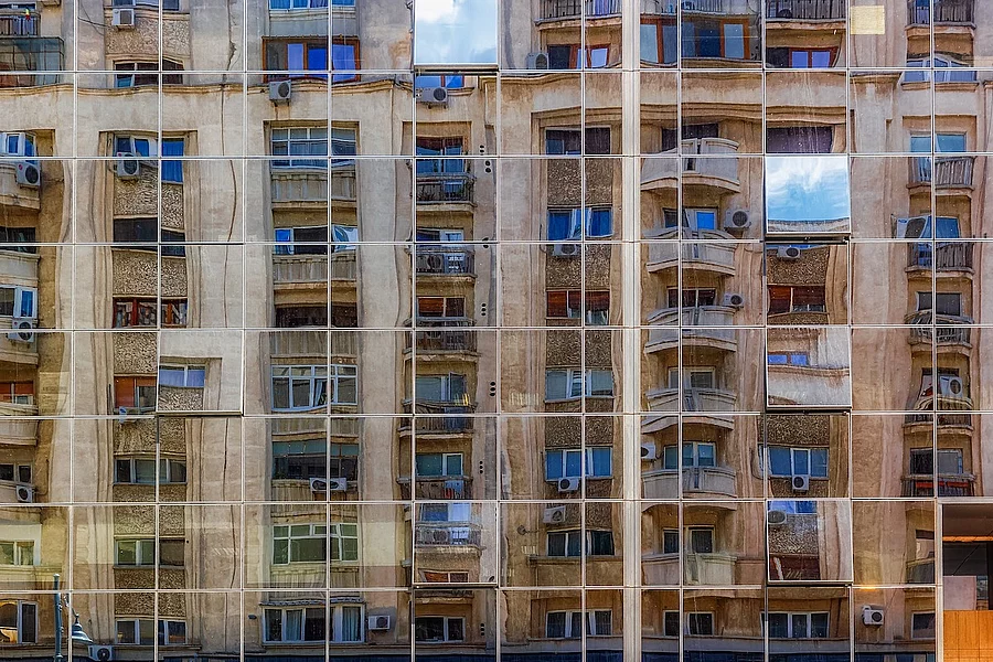 Fotografia prezintă fațada din sticlă reflectorizantă a unei clădiri moderne din București, în care se oglindesc clădiri rezidențiale mai vechi, în stil clasic.