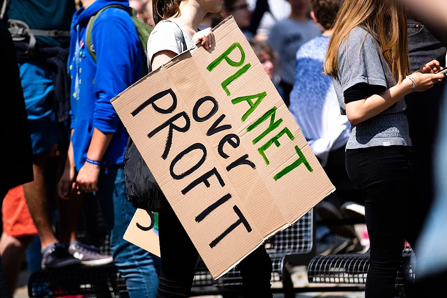 În imagine se vede o persoană care ține un carton cu mesajul „PLANETA înaintea PROFITULUI” în cadrul unei manifestații.