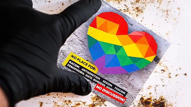 A hand wearing a black glove is holding a sticker featuring a colourful rainbow heart and the slogan ‘NO PLACE FOR: RACISM... NO DISCUSSION!’ to highlight the fight against discrimination.